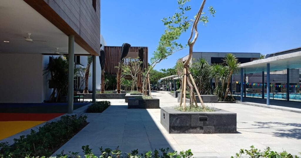 Modern ais indonesia bali campus courtyard with trees, open pathways, and swimming pool under bright blue sky.