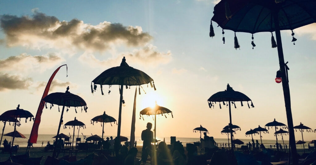 Tropical beach at sunset with silhouettes and traditional umbrellas, highlighting regional differences and local rules for alcohol across indonesia.
