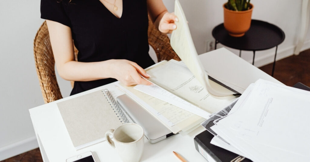 Osoba sortująca wydrukowane dokumenty podróżne i formularze na biurku obok laptopa i kawy, przygotowująca papierkową robotę na wycieczkę na Bali.