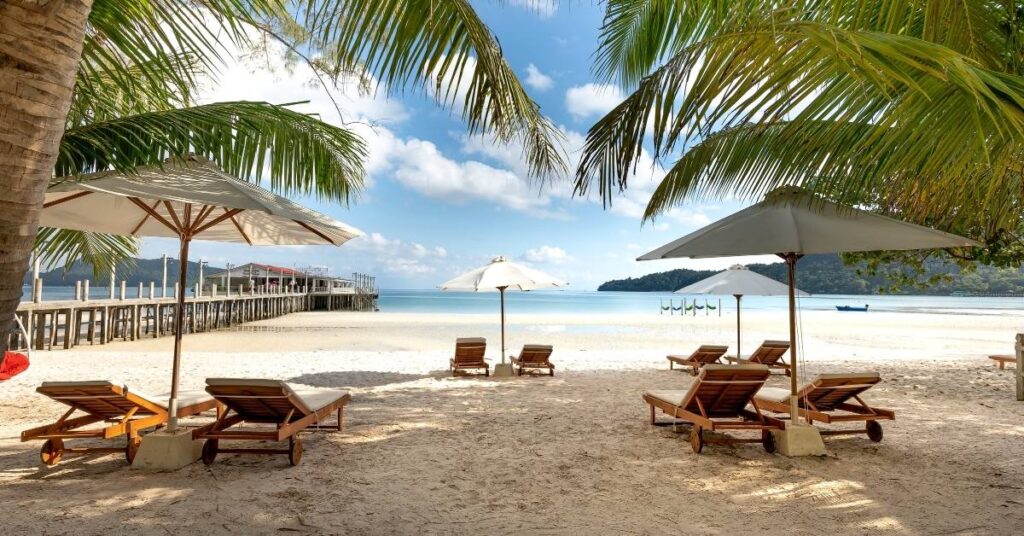 Тихий тропічний пляж на Балі з дерев'яними шезлонгами і білими парасольками під пальмами з видом на спокійну бірюзову воду і дерев'яний пірс. Ілюстрація до статті про безпеку подорожей "Балі - це безпечно". "