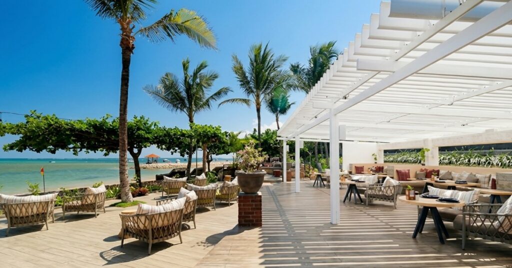 Beachfront café in sanur with outdoor seating, palm trees, and ocean views.
