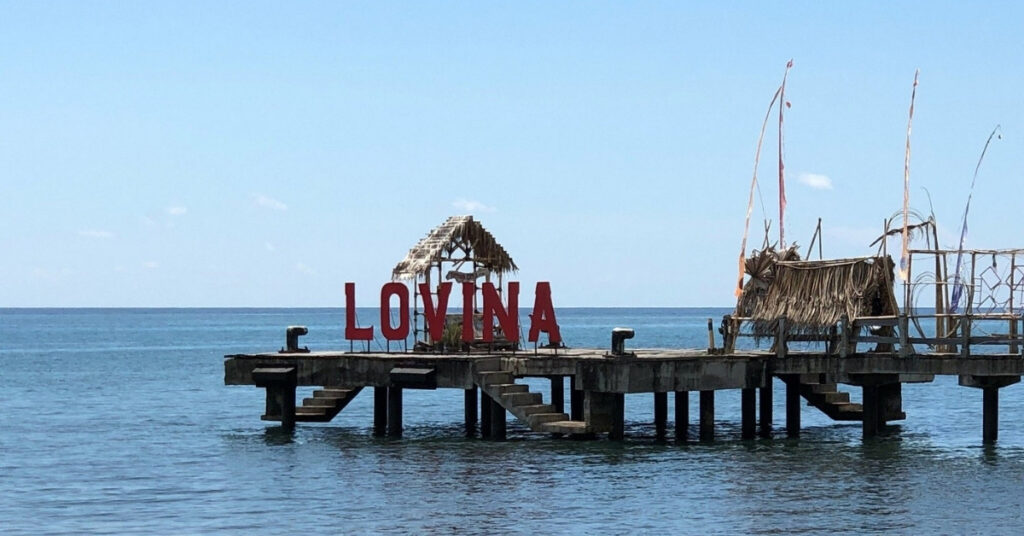 Lovina pier with red “Lovina” sign over calm north-coast waters. Slow, quiet pace