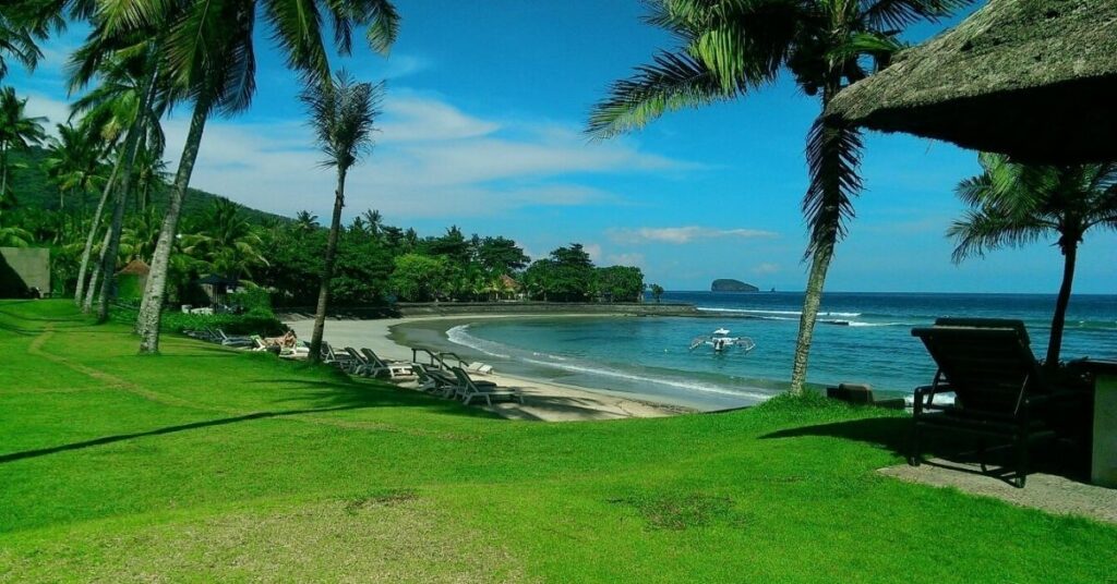 Palm-lined lawn with sun loungers facing a calm bay in Candidasa, East Bali. Black-sand beach, jukung boat, and a quiet, laid-back setting for retirees.