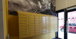 “「オフィスのロビーにずらりと並んだ黄色い郵便受け。組織的な郵便物の取り扱いと、バーチャルオフィスの住所として安全な郵送先を示している。.