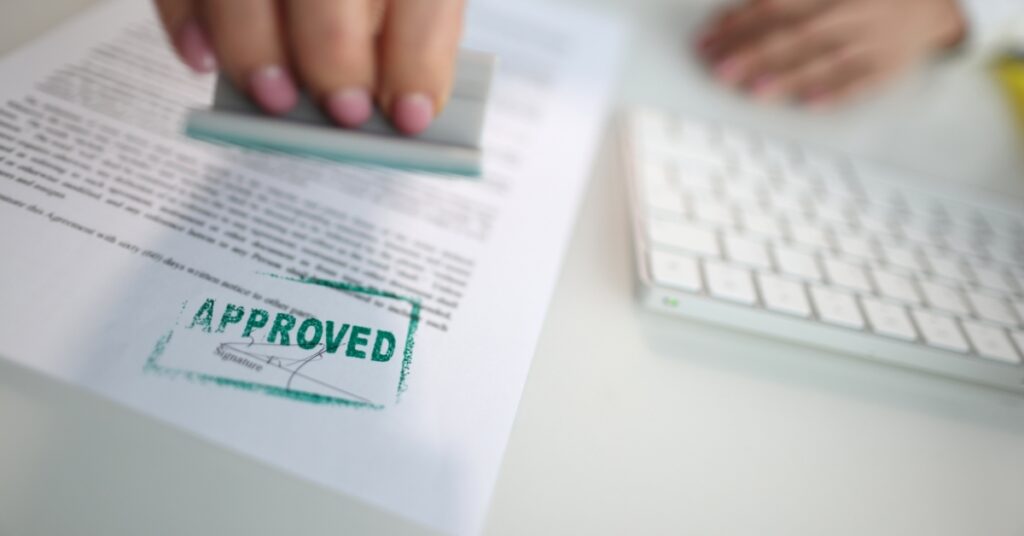 Håndstempling av et "godkjent"-merke på et dokument ved siden av et tastatur, som symboliserer at visumet eller tillatelsen er godkjent etter sponsorbytte.