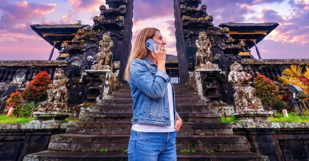 Tourist using a smartphone in front of a balinese temple gate. Staying connected in bali with a travel esim plan from visa-indonesia. Com