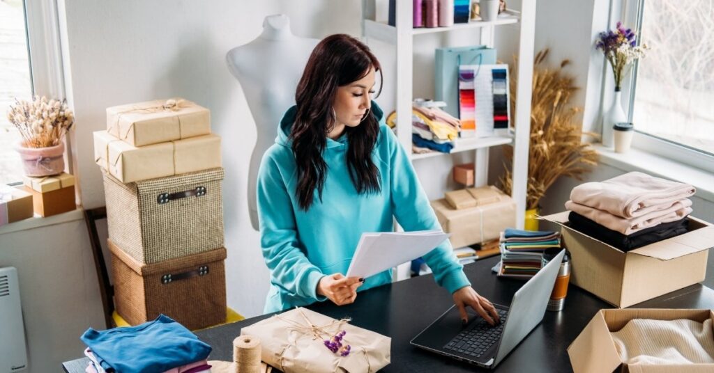 Woman running a small product business, checking documents and laptop in a studio filled with boxes and fabrics, illustrating small enterprises in indonesia.