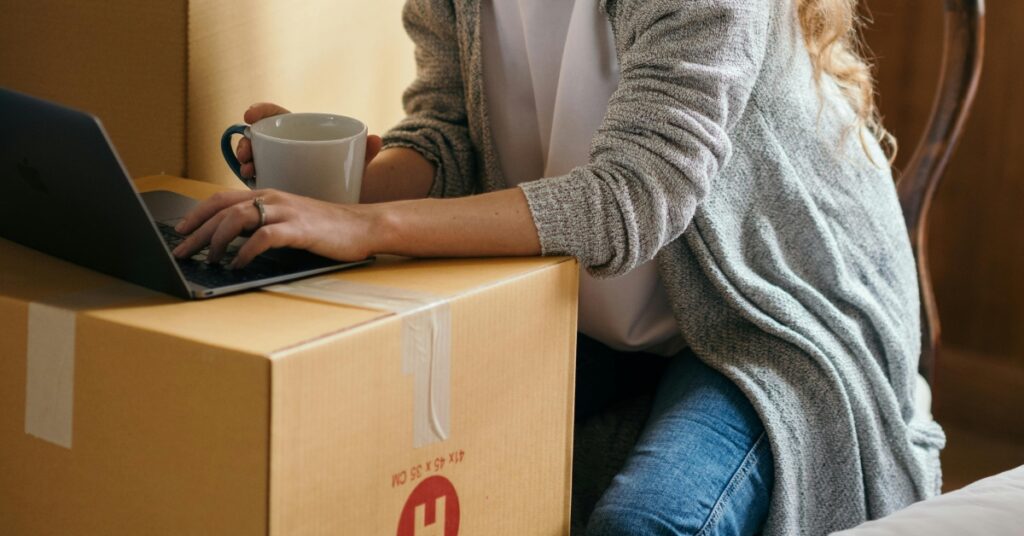 Жінка користується ноутбуком, тримаючи в руках чашку з кавою, сидячи на картонній коробці під час переїзду додому, подаючи заяву на отримання візи на друге місце проживання в Індонезії