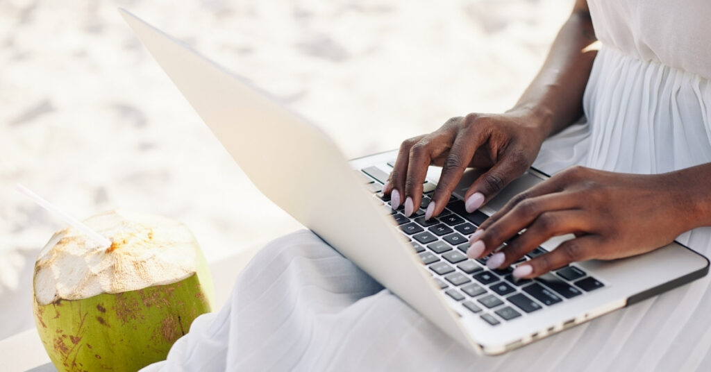 Hands typing on a laptop beside a fresh coconut on a sandy beach, showing an online indonesia gci (global citizenship) application vibe.