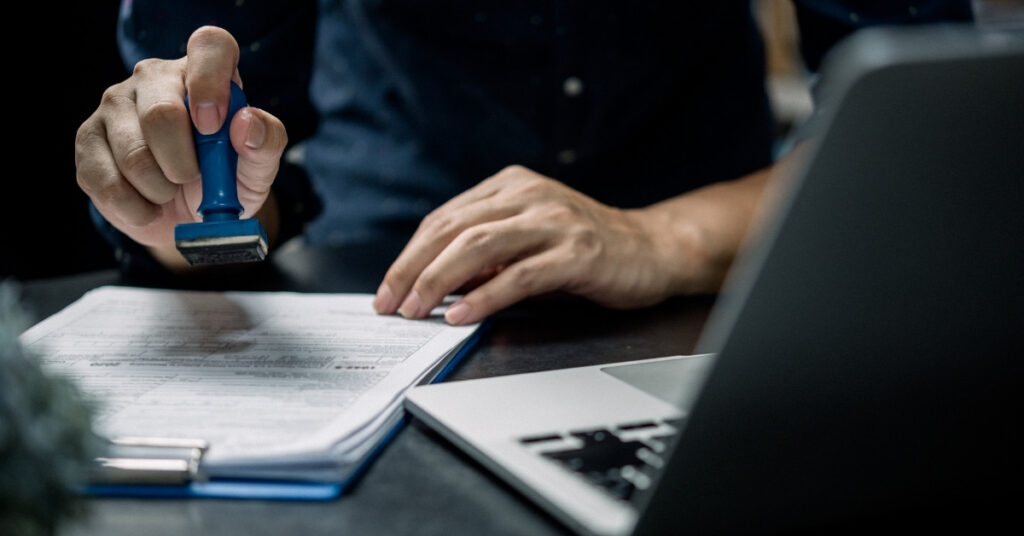 Mano che tiene un timbro di gomma su un documento accanto a un computer portatile, suggerendo di controllare i documenti di immigrazione per evitare un soggiorno eccessivo in Indonesia.