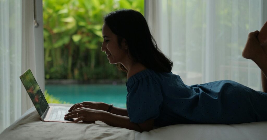 Lachende vrouw met laptop in slaapkamer van villa met uitzicht op privézwembad en tropische tuin, levensstijl op afstand werken in Bali.