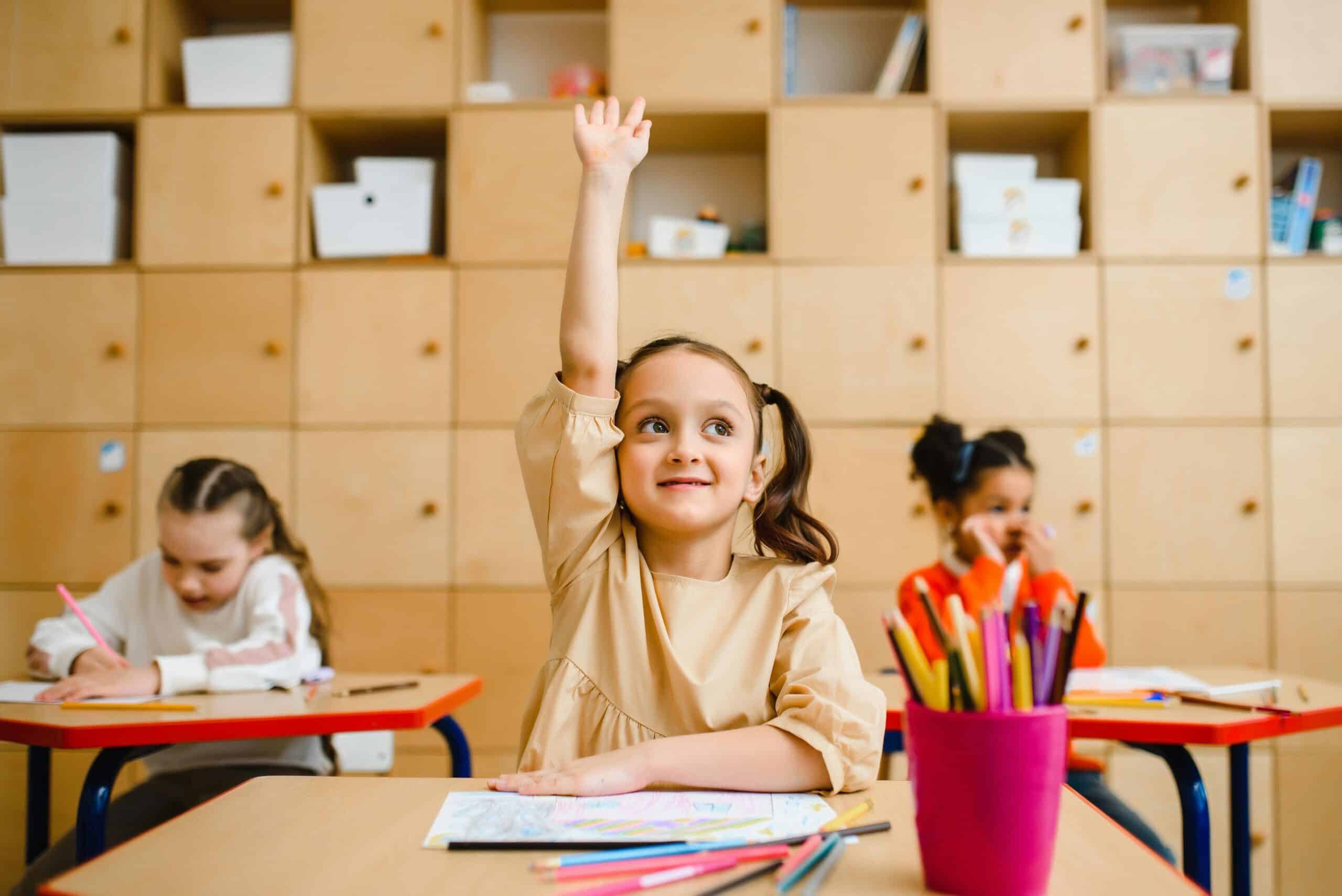 A cheerful young girl raising her hand in a classroom, with colorful pencils and classmates in the background.