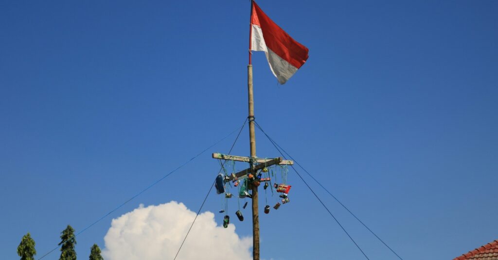 Alto poste de palma areca con premios en la parte superior y la bandera indonesia ondeando en lo alto, bajo un cielo azul brillante durante un evento tradicional de Panjat Pinang.