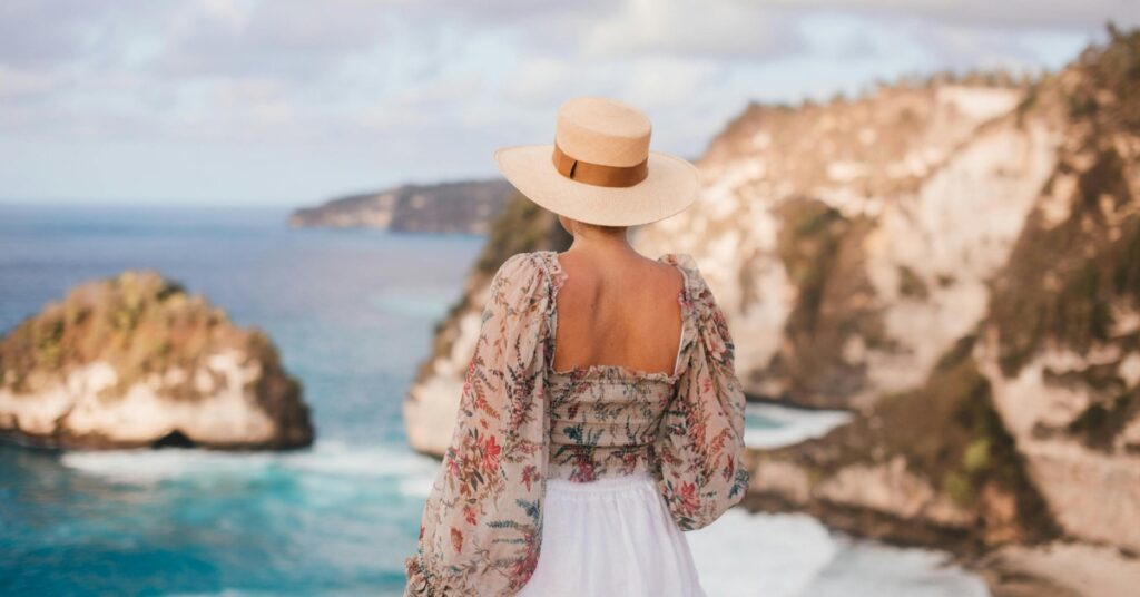 Женщина в соломенной шляпе и цветочной блузке стоит на смотровой площадке у скалы, любуясь бирюзовыми водами и изрезанными прибрежными скалами в Бали, Индонезия.
