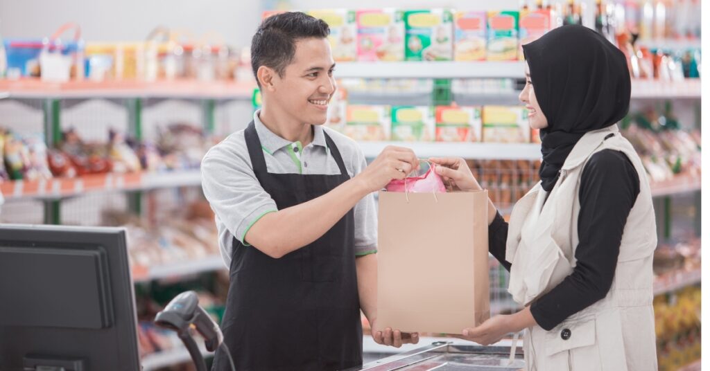 En kassörska ger en shoppingväska till en leende kvinna i hijab på en stormarknad, som representerar halalcertifierade konsumentprodukter.