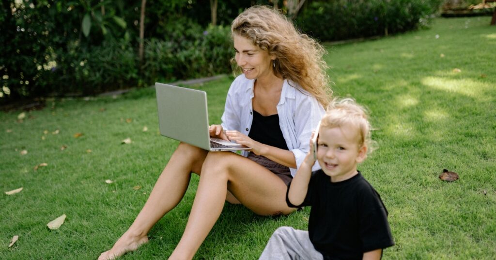 Мати працює за ноутбуком на свіжому повітрі з маленькою дитиною, яка грається на траві поруч в Індонезії