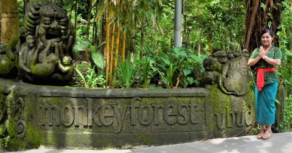Entrée de la forêt des singes à ubud avec une femme locale en costume traditionnel accueillant les visiteurs à côté du panneau en pierre sculptée.