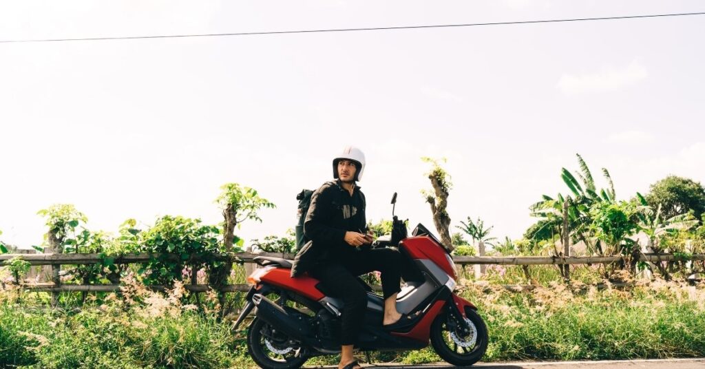 Турист на червоному скутері в балі, в шоломі і з рюкзаком, припаркований вздовж мальовничої сільської дороги.