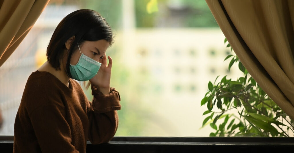 Worried woman wearing a face mask sits by a window, symbolizing stress while seeking legal help or filing an objection to a deportation order.