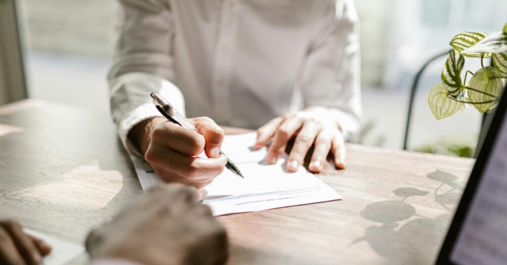 Persona che firma documenti ufficiali rptka su una scrivania di legno illuminata dalla luce del sole.