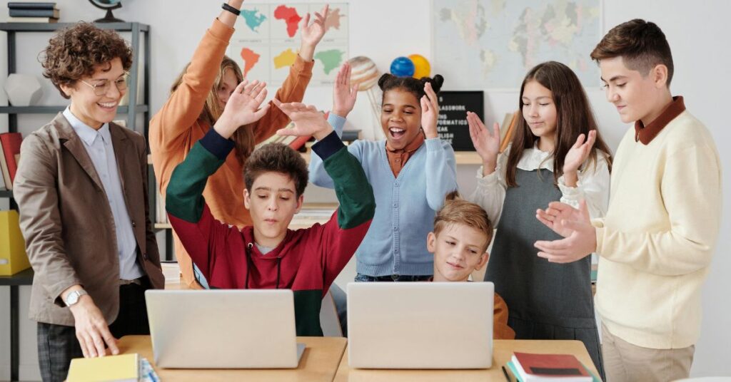 チームワークと学習成果の成功を象徴する、ノートパソコンを囲んで祝う多様な生徒と教師のグループ。