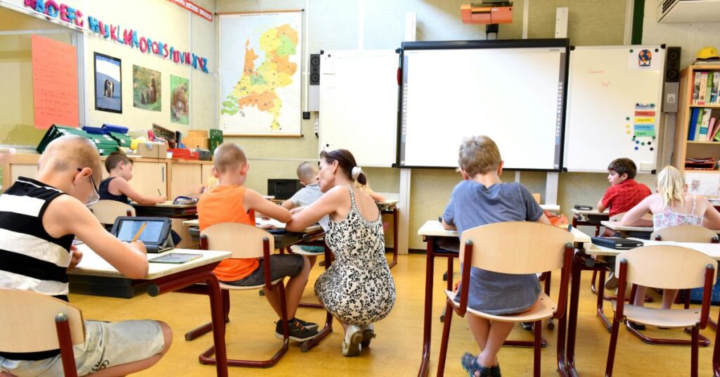 アルファベットのウォールアートを背景に、明るい教室で若い生徒を指導する教師。