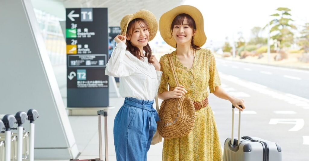 To smilende kvinnelige turister med kofferter utenfor en flyplassterminal, klare til å reise inn i Indonesia med visum ved ankomst eller visumfri innreise.