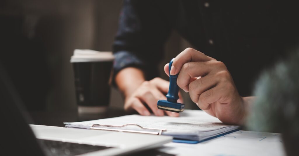 Gros plan d'une personne apposant un tampon sur des documents de visa approuvés, à côté d'une tasse de café et d'un ordinateur portable, symbolisant la vérification professionnelle d'un agent de visa.