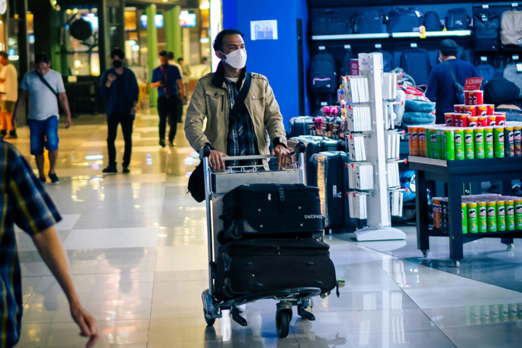 Чоловік у масці штовхає візок з багажем у залі прибуття аеропорту Балі, проходячи повз магазин безмитної торгівлі та стійки реєстрації багажу.