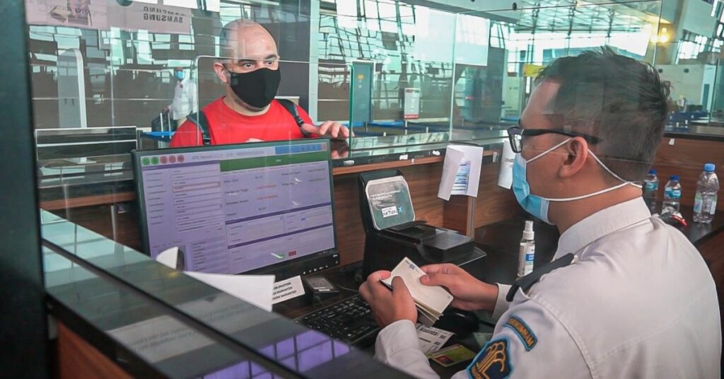 Un funzionario dell'immigrazione controlla un passaporto allo sportello di un aeroporto mentre un viaggiatore parla attraverso il vetro, illustrando cosa fare dopo un ritardo.