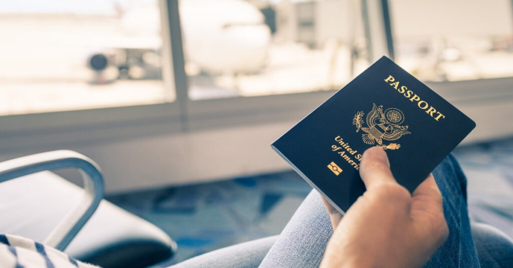 Hand holding a u. S. Passport in an airport lounge with an aircraft outside the window, symbolizing foreign nationals departing indonesia.